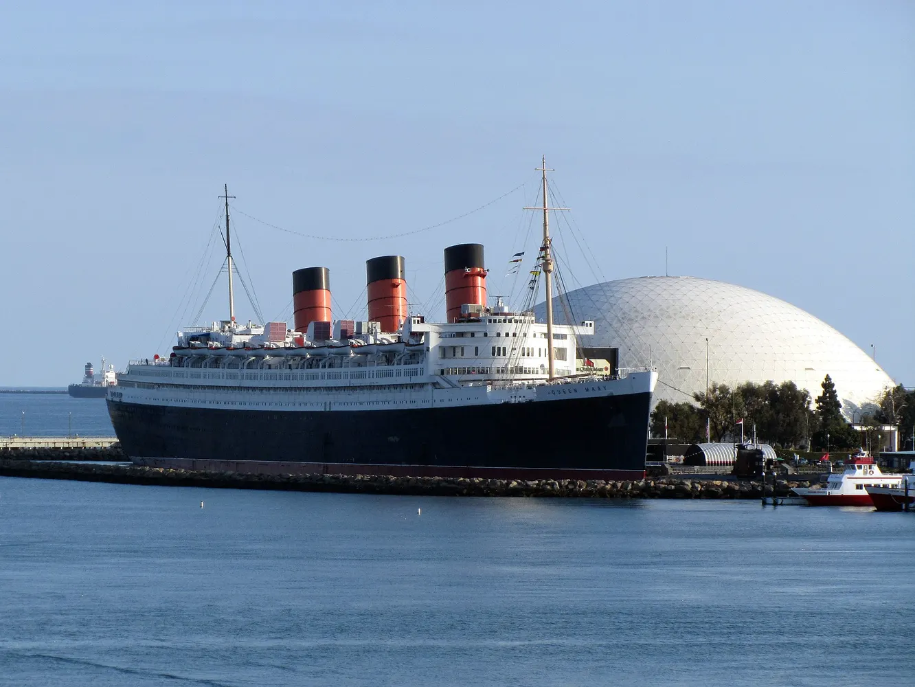 The Queen Mary ocean liner docked at Long Beach with the white dome behind it under a clear sky.