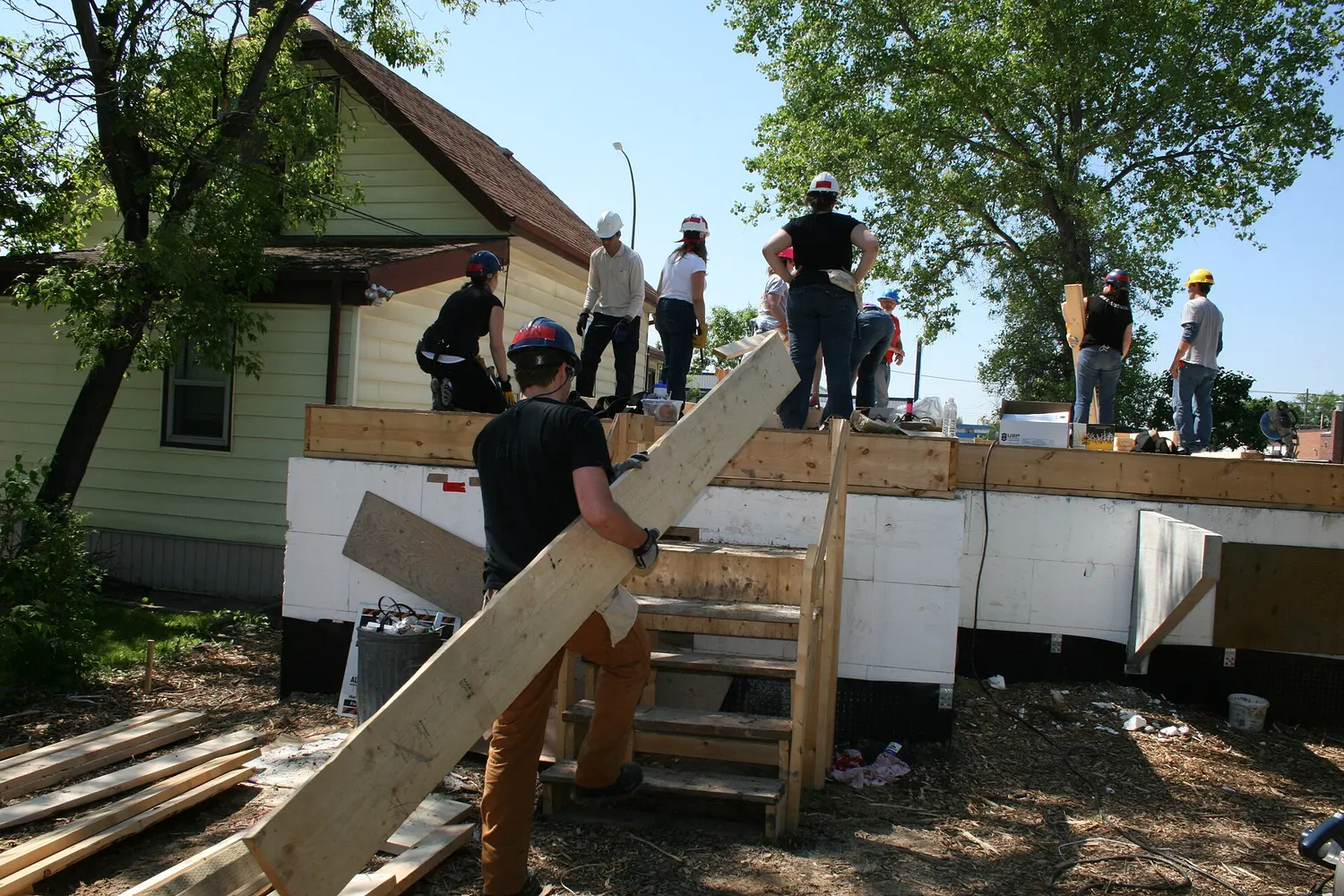 Volunteers in hard hats raise wall framing on a Habitat for Humanity house build on a sunny day.