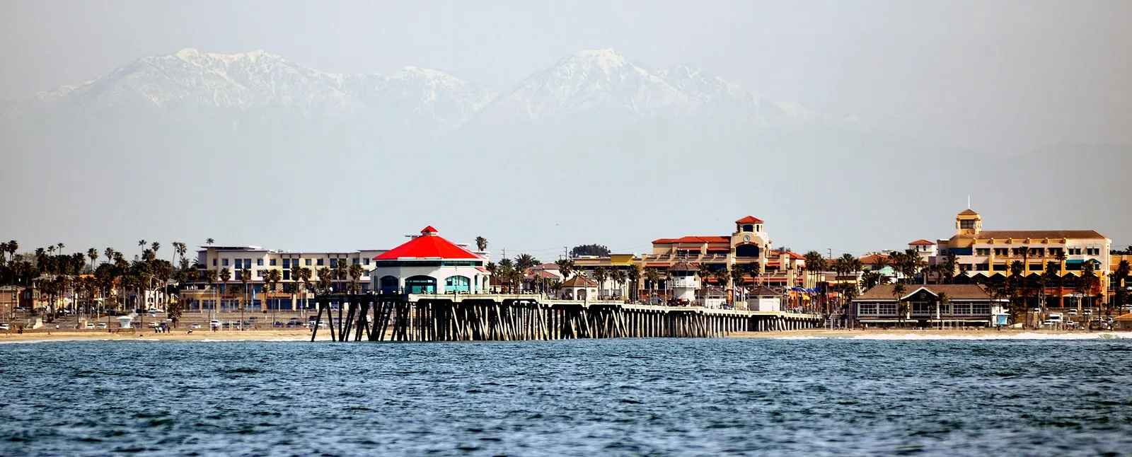 The Huntington Beach pier stretches over the surf with the town and snow-capped mountains behind it.