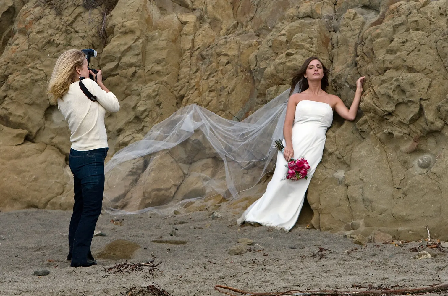 A wedding photographer lines up a shot of a bride posing against coastal rocks.