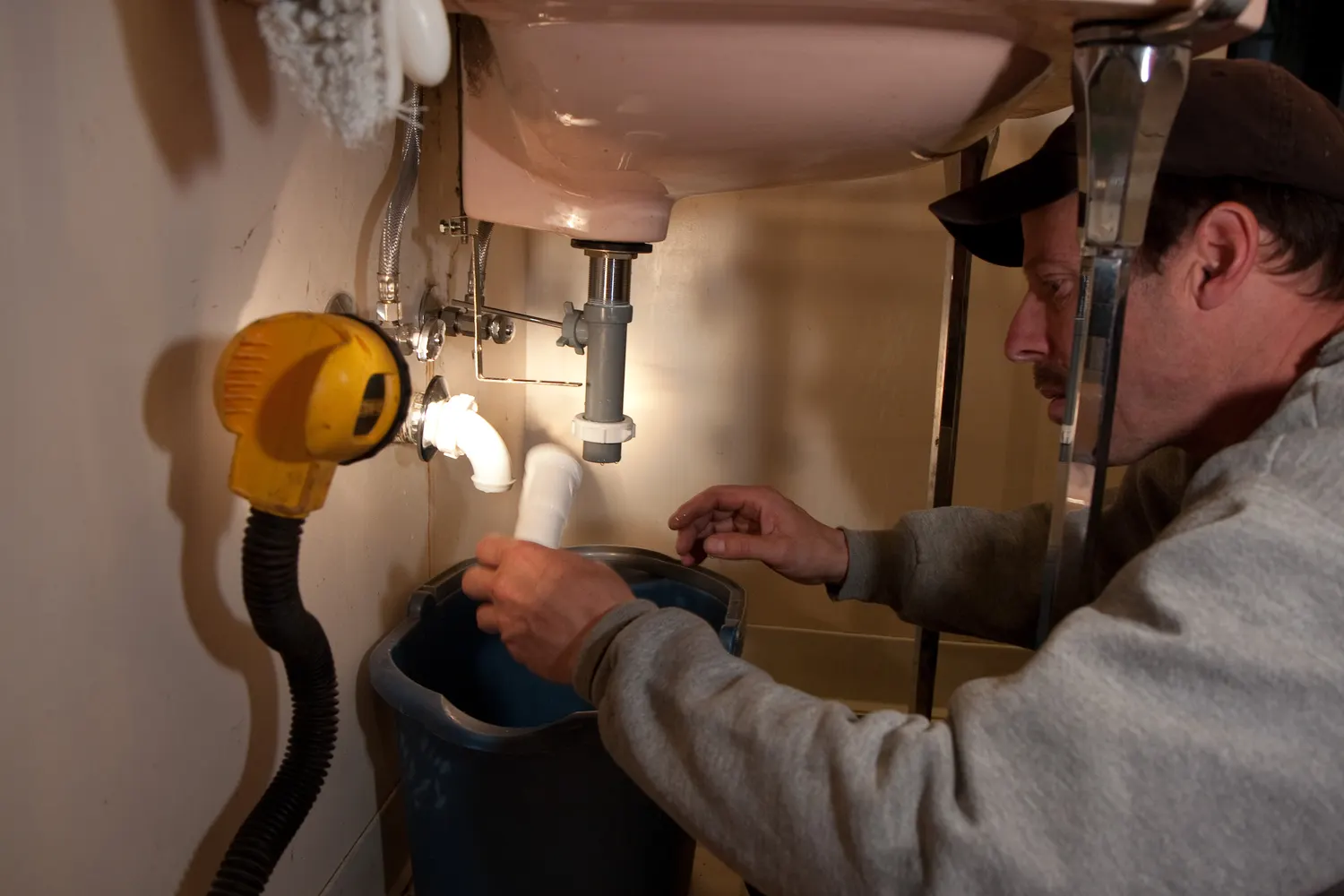 A plumber kneels under a kitchen sink and works on the drain pipes.