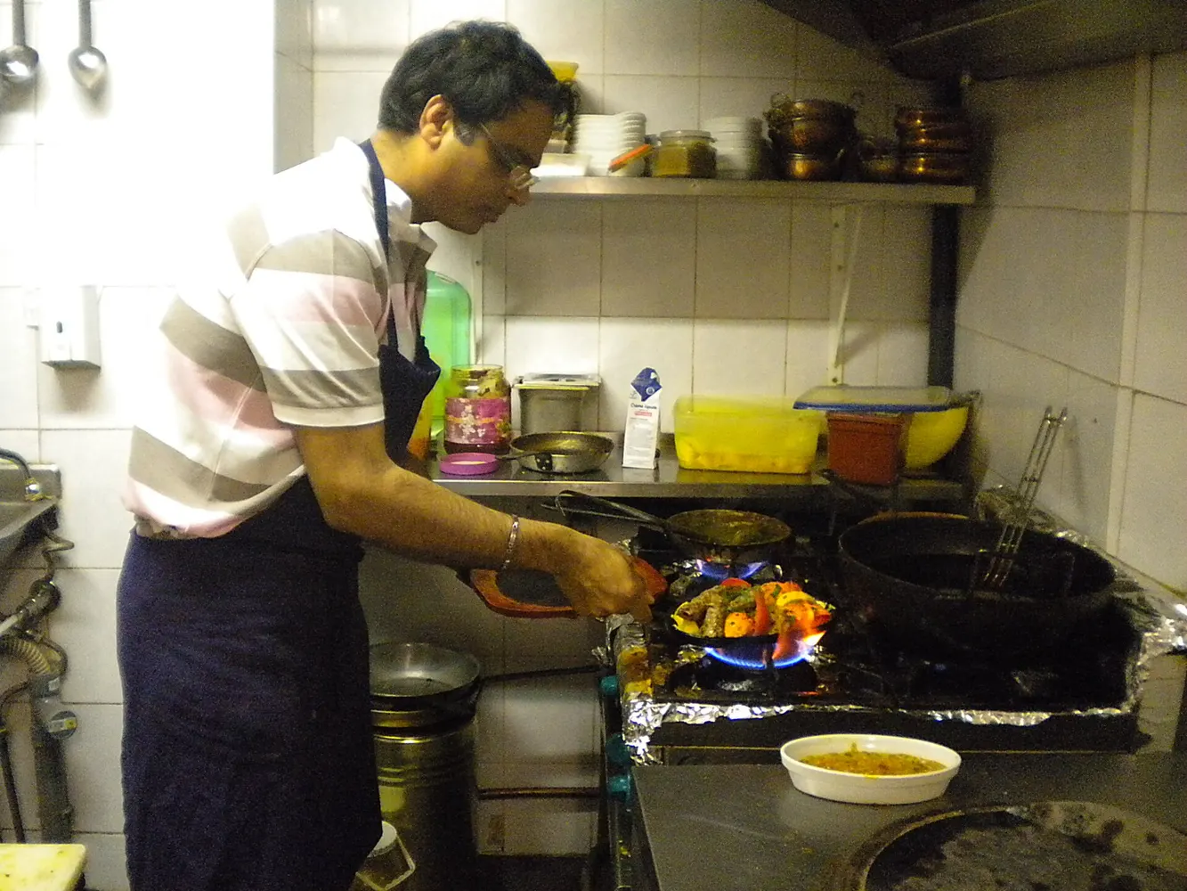 A cook works a wok over an open flame on a restaurant kitchen stove.
